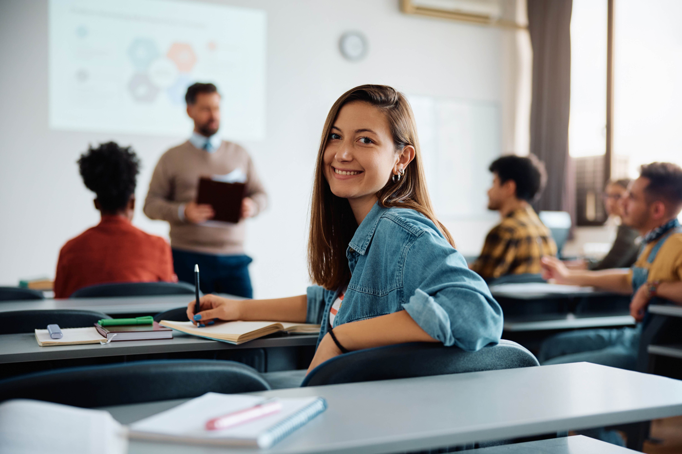 Junge Frau bei einem Kurs in einem Seminarraum mit anderen Teilnehmenden und Vortragendem im Hintergrund